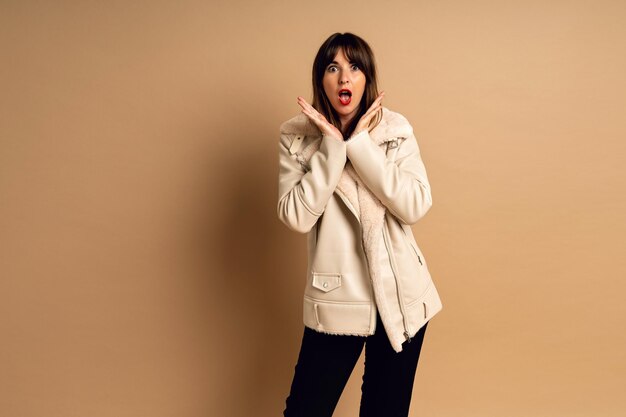 Elegante mujer morena joven posando en abrigo de piel, fondo de estudio beige, horario de invierno.