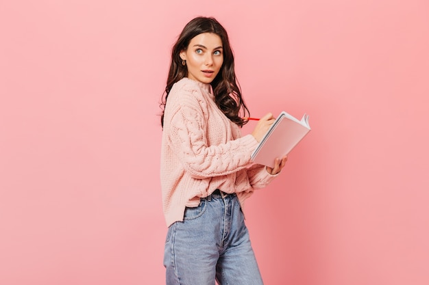 Elegante mujer morena rizada posando con cuaderno y bolígrafo rojo sobre fondo rosa.