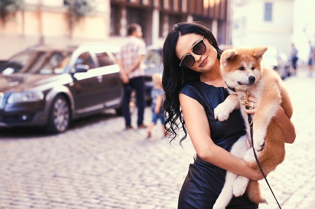 Una elegante mujer morena con un vestido negro y gafas de sol, parada en una calle y sosteniendo un lindo cachorro Akito.