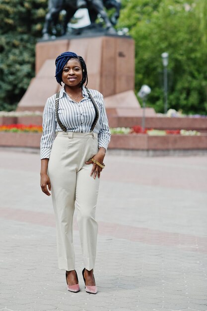 Elegante mujer de negocios afroamericana en pantalones con tirantes y blusa posada al aire libre