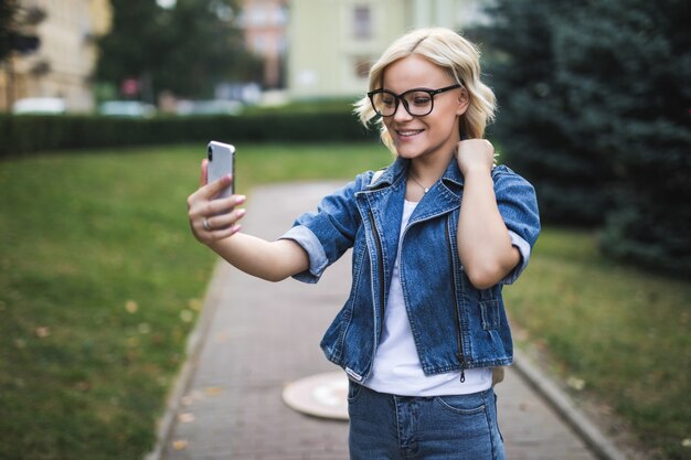 Elegante mujer de niña rubia de moda bonita feliz en jeans suite hace selfie en su teléfono en la ciudad por la mañana