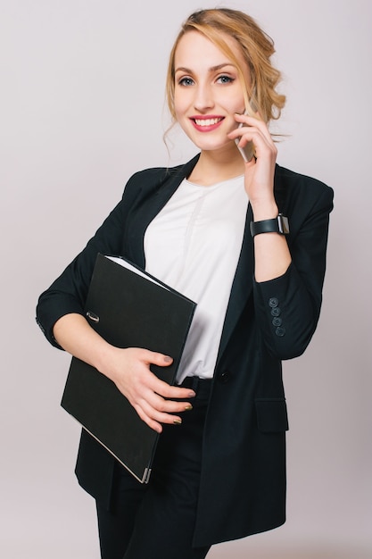 Elegante mujer de oficina joven de moda en traje, sosteniendo la carpeta, hablando por teléfono aislado. Estado de ánimo alegre, éxito, carrera, estar ocupado, trabajar, verdaderas emociones positivas