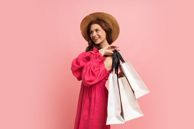 Elegante mujer pelirroja con bolsas blancas posando en vestido de gravamen rosa con mangas sobre pared rosa.