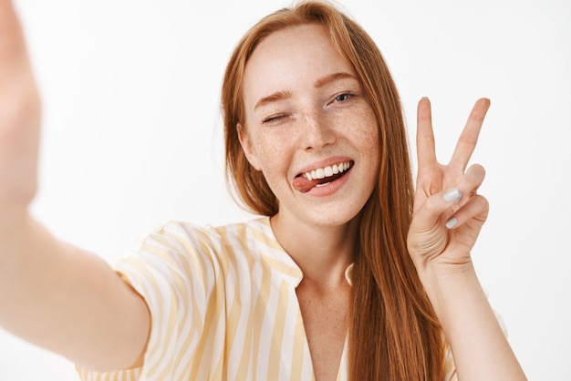 Elegante mujer pelirroja hermosa con lindas pecas sacando la lengua y sonriendo de alegría guiñando un ojo mostrando felizmente el signo de la paz, tomando selfie en smartphone