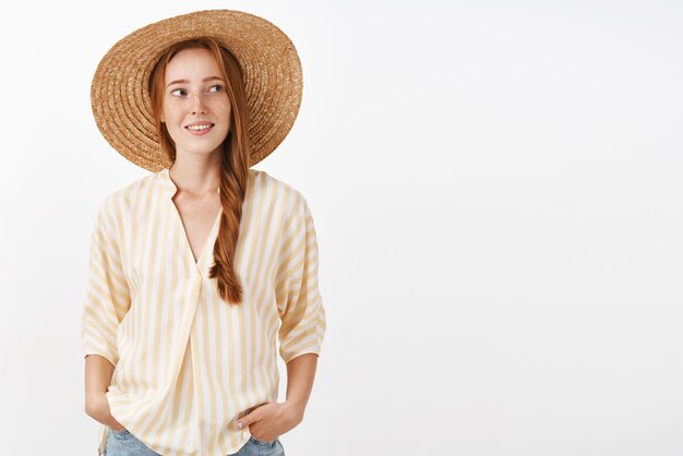 Elegante mujer pelirroja que va a la playa con sombrero de paja para no broncearse girando a la derecha con expresión feliz y despreocupada tomados de la mano en los bolsillos disfrutando de un cálido día soleado de verano