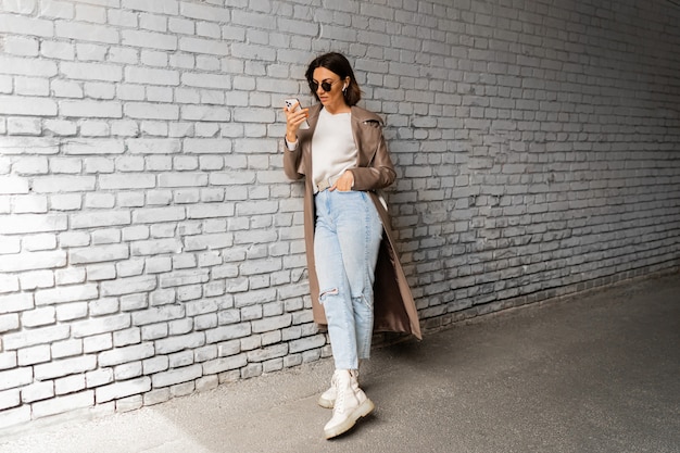 Elegante mujer de pelo corto con auriculares en abrigo de cuero casual y gafas de sol con smartphone y posando sobre pared de ladrillo urbano