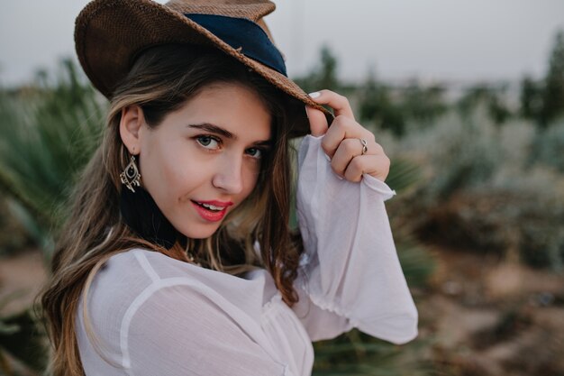 Elegante mujer de pelo largo con elegante sombrero se ve, mientras camina en el hermoso parque exótico. Retrato de primer plano de una mujer joven y bonita en aretes de moda y camisa posando con expresión facial enigmática