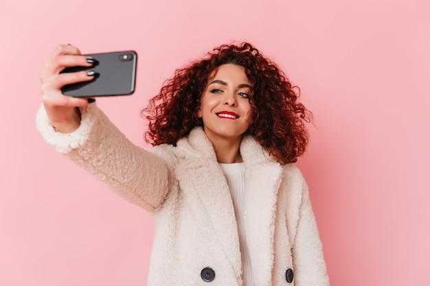 Elegante mujer de pelo oscuro rizado con abrigo de piel ecológica se muerde el labio y toma selfie en espacio rosa.