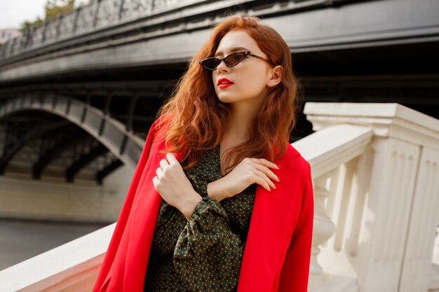 Elegante mujer con pelos de jengibre ondulado posando al aire libre en la chaqueta roja.