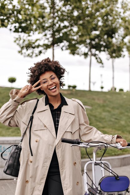 Elegante mujer rizada de piel oscura muestra el signo de la paz afuera Dama activa en gabardina beige posa con bicicleta afuera