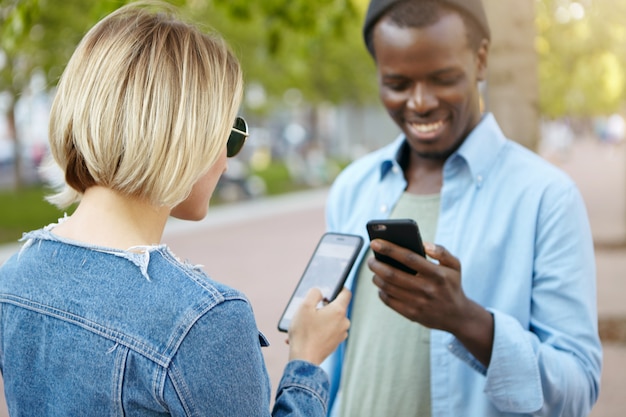 Elegante mujer rubia con chaqueta de mezclilla y gafas de sol reuniéndose con su amigo africano en la calle, sosteniendo teléfonos celulares en las manos, intercambiando sus números de teléfono para mantener sus relaciones