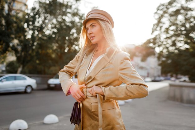 Elegante mujer rubia con gorra beige y chaqueta caminando por la calle. Mirada otoñal. Luz del atardecer. Monedero elegante.
