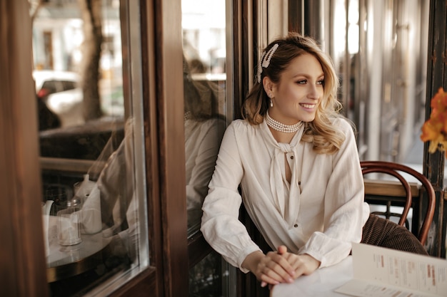 Elegante mujer rubia hermosa en blusa blanca elegante, joyas de perlas sonríe ampliamente, mira hacia otro lado y se sienta junto a la mesita en el café de la calle