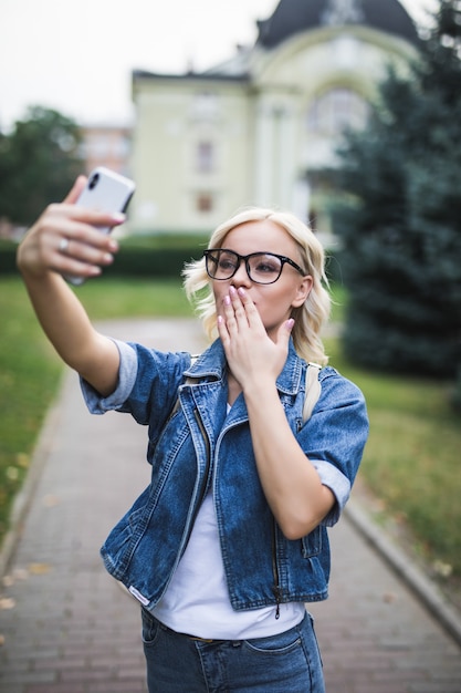 Elegante mujer rubia de moda en jeans suite hace selfie dar besos en su teléfono en la ciudad por la mañana