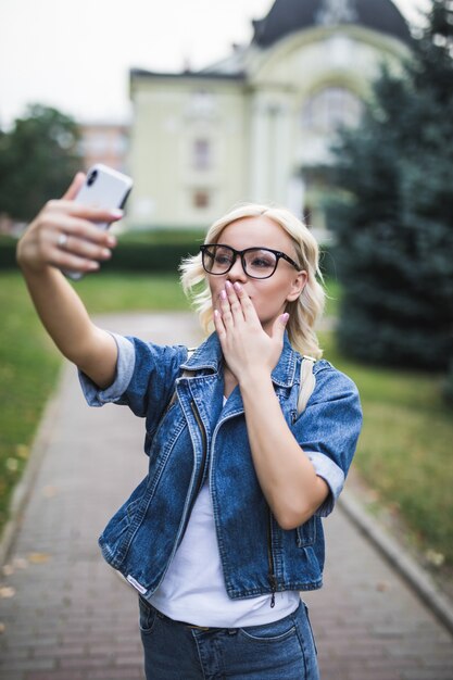 Elegante mujer rubia de moda en jeans suite hace selfie dar besos en su teléfono en la ciudad por la mañana