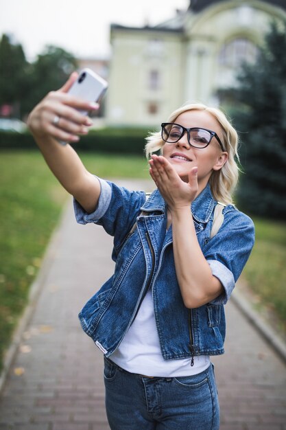 Elegante mujer rubia de moda en jeans suite hace selfie dar besos en su teléfono en la ciudad por la mañana