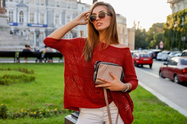 Elegante mujer rubia en moda otoño ourfit posando en la calle. Llevaba gafas de sol, jersey y jeanse blanco.