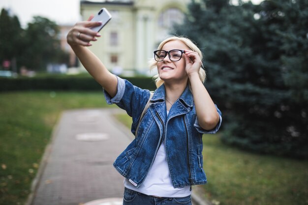 Elegante mujer rubia de moda sonriente en suite de jeans hace selfie en su teléfono en la ciudad por la mañana