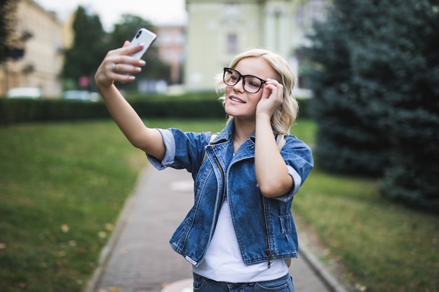 Elegante mujer rubia de moda sonriente en suite de jeans hace selfie en su teléfono en la ciudad por la mañana