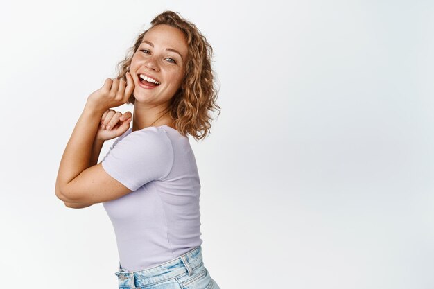 Elegante mujer rubia con peinado corto y rizado, tocando su brillante piel facial saludable, sonriendo a la cámara, de pie sobre fondo blanco.