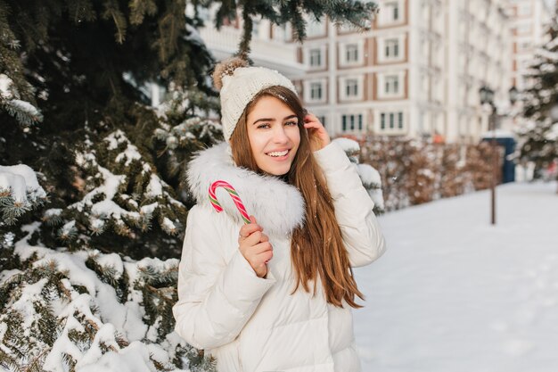 Elegante mujer rubia posando con sonrisa feliz comiendo bastón de caramelo dulce en día de invierno. Retrato de hermosa mujer europea con gorro de punto de pie junto al abeto nevado y riendo.