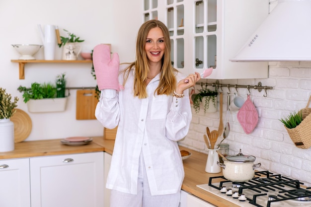 Elegante mujer rubia posando en su linda cocina con pijama de lino blanco.