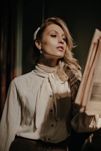 Elegante mujer rubia segura de sí misma en blusa blanca y pantalón marrón lee el periódico y posa en el pasillo oscuro