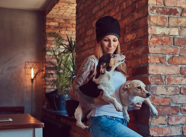 Una elegante mujer rubia tatuada con camiseta y jeans sostiene dos lindos perros.