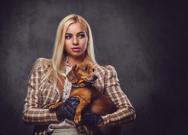 Una elegante mujer rubia vestida con una chaqueta antigua sostiene un perro tejón rojo.