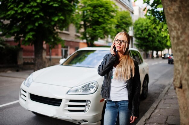 Elegante mujer rubia vestida con gafas de jeans y chaqueta de cuero con teléfono móvil contra un automóvil de lujo Retrato de modelo urbano de moda