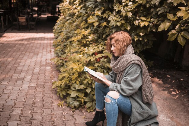 Elegante mujer sentada a bordo y atentamente leyendo el libro cerca de la planta