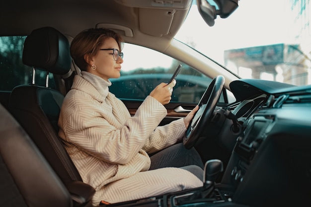 Elegante mujer sentada en el coche vestida con abrigo estilo de invierno y gafas con smartphone