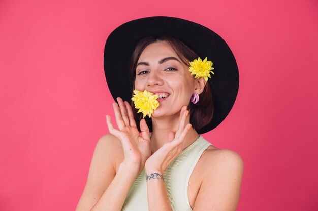 Elegante mujer con sombrero, sonriendo con dos ásteres amarillos, lindo sostener una flor en la boca, humor primaveral, emociones felices espacio aislado