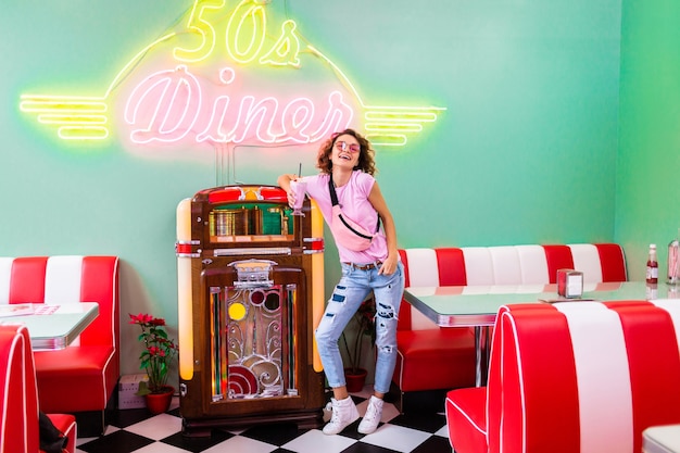 Foto gratuita elegante mujer sonriente atractiva en café retro de los años 50 en la máquina de discos de música bebiendo cócteles de batido de leche en traje de hipster divirtiéndose riendo en un estado de ánimo alegre