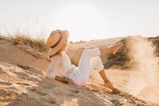 Elegante mujer sonriente atractiva feliz posando en el desierto vestida con ropa blanca con sombrero de paja y gafas de sol en la puesta de sol