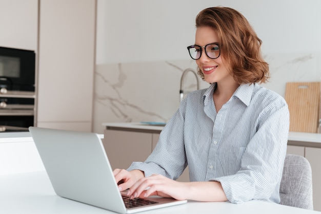 Elegante mujer sonriente en gafas y camisa a rayas usando la computadora portátil mientras se sienta a la mesa en la cocina