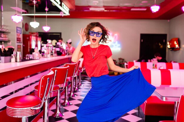 Elegante mujer sonriente con traje colorido en un café retro de los años 50 bailando con una falda azul chaqueta y gafas de sol de camisa roja divirtiéndose de buen humor