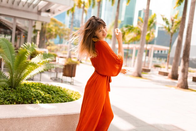 Elegante mujer en vestido naranja posando en el paseo marítimo con palmeras y rascacielos en la gran ciudad moderna