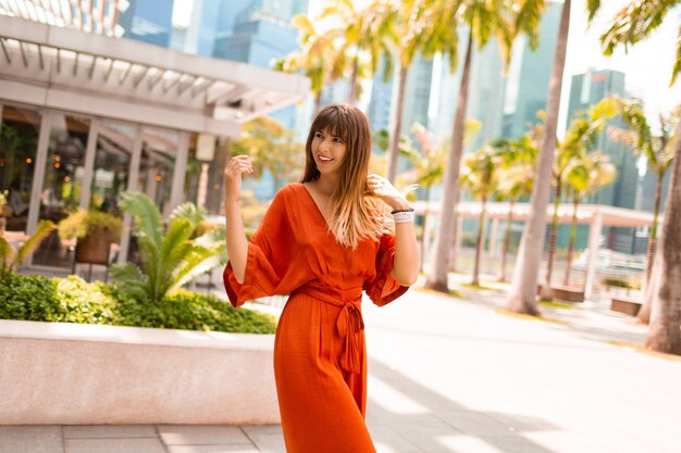 Elegante mujer en vestido naranja posando en el paseo marítimo con palmeras y rascacielos en la gran ciudad moderna