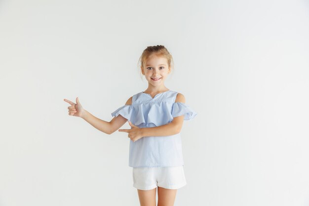 Elegante niña sonriente posando en ropa casual aislado sobre fondo blanco de estudio. Modelo de mujer rubia caucásica. Emociones humanas, expresión facial, infancia. Apuntando a la barra espaciadora vacía.