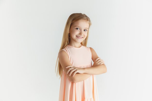 Elegante niña sonriente posando en vestido aislado sobre fondo blanco de estudio. Modelo de mujer rubia caucásica. Las emociones humanas, la expresión facial, la infancia. De pie con las manos cruzadas.