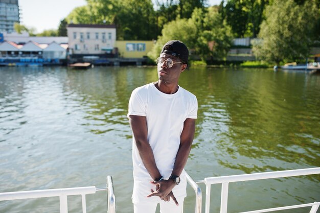 Elegante niño afroamericano vestido con ropa blanca, gafas y gorra contra el muelle del lago Moda callejera de jóvenes negros