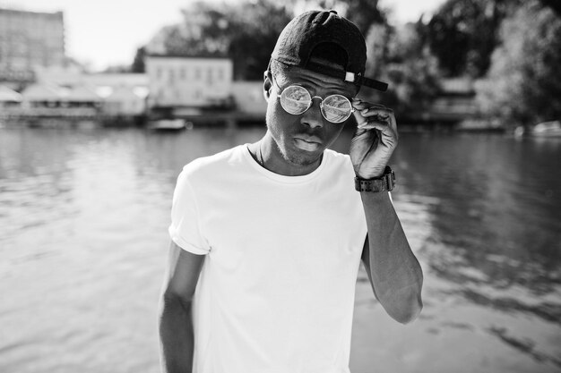 Elegante niño afroamericano vestido con ropa blanca, gafas y gorra contra el muelle del lago Moda callejera de jóvenes negros