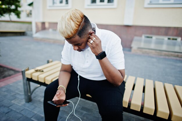 Elegante niño musulmán árabe con cabello originalmente posado en las calles sentado en un banco y escuchando música desde los auriculares del teléfono