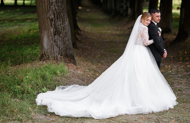 Elegante novia en vestido de novia de pie detrás del guapo novio en el jardín