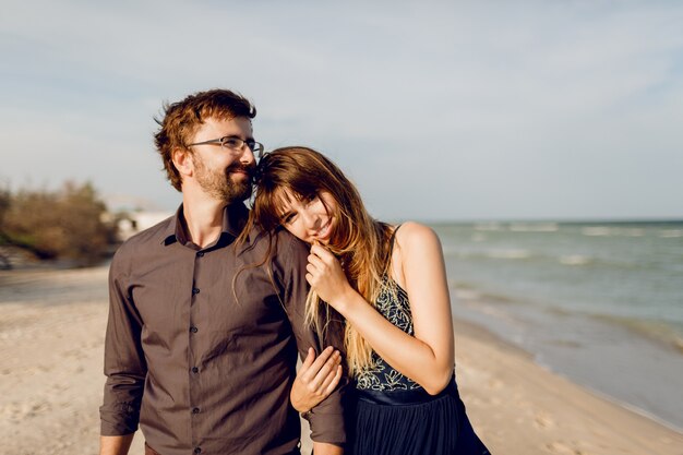 Elegante pareja de enamorados caminando en la playa soleada tarde, mujer feliz avergonzando a su marido.