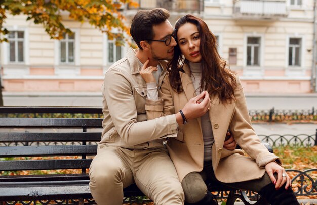 Elegante pareja de enamorados posando al aire libre. Tendencias de la moda de otoño. Modelo morena con chico elegante abrigo beige sentado en el banco.