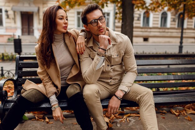 Elegante pareja de enamorados posando al aire libre. Tendencias de la moda de otoño. Modelo morena con chico elegante abrigo beige sentado en el banco.
