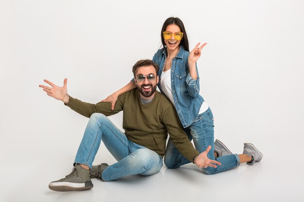 Elegante pareja sentada en el piso aislado, mujer y hombre muy sonrientes en jeans, con gafas de sol, divirtiéndose juntos, extendiendo las manos en emoción positiva