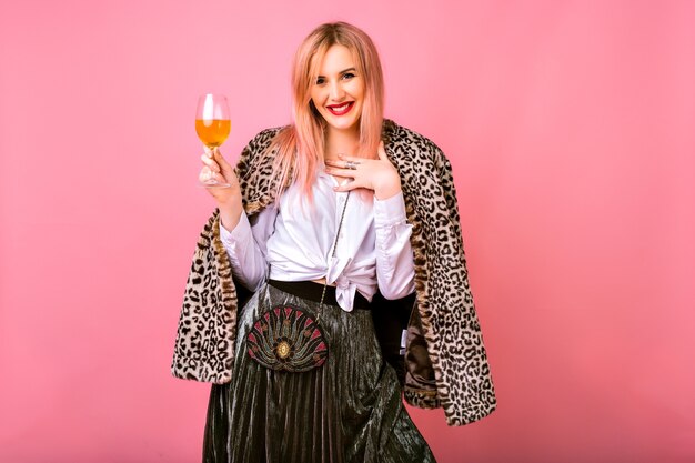 Elegante y positiva mujer joven y bonita divirtiéndose, vistiendo traje de cóctel brillante de noche y abrigo de moda con estampado de leopardo de piel, fondo rosa, disfrutando de la fiesta de vacaciones de invierno